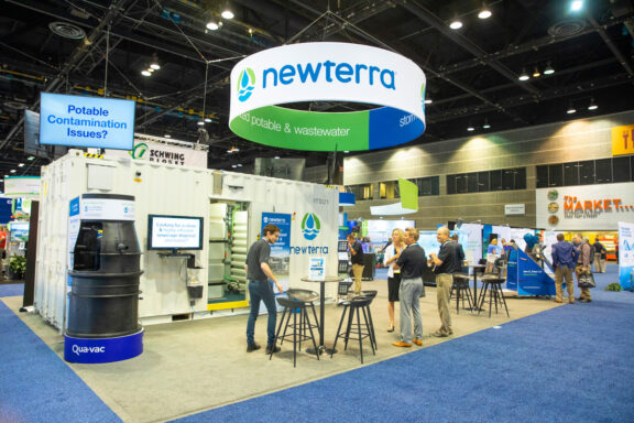 Newterra's professional vendor booth at an industry event with potential customers and Newterra employees. Circular banner attached above the open floorplan with banners covering the walls with Newterra branded information and a product demonstration.