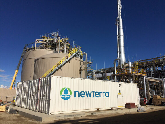 Modular water treatment technology contained within a Newterra branded shipping container with large beige storage tanks behind four modular units
