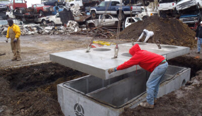 Technician in red hoodie guides the concrete lid of a water treatment technology at a automotive junkyard to its base container buried in the group with the aid of a crane, cars for crushing are stacked in the background