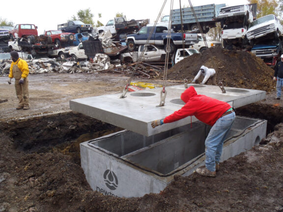 Technician in red hoodie guides the concrete lid of a water treatment technology at a automotive junkyard to its base container buried in the group with the aid of a crane, cars for crushing are stacked in the background
