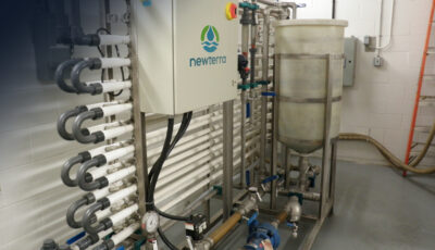 Newterra ultrafiltration system mounted on a metal wall with a series of grey tubes covered in white plastic tubing, upper left corner contains a black shadow from where the camera lens was covered during the shooting of the image.