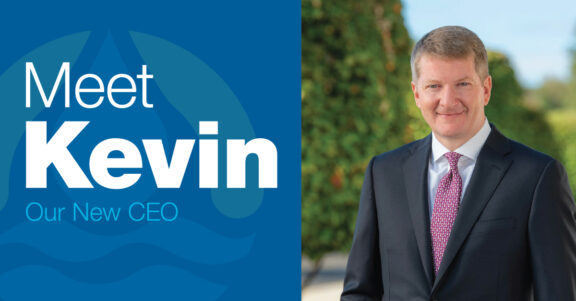 Article banner of split image of Newterra's CEO, Kevin on the right wearing a black suit, white shirt, and multicolor patterned tie with white text containing Meet Kevin, our New CEO on the left in front of a background containing a subtle underlay of the Newterra logo on a blue background underneath the white text