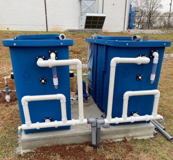 Two blue number four plastic containers containing Newterra water treatment technology sit on a concrete slab. The containers have lids and 2