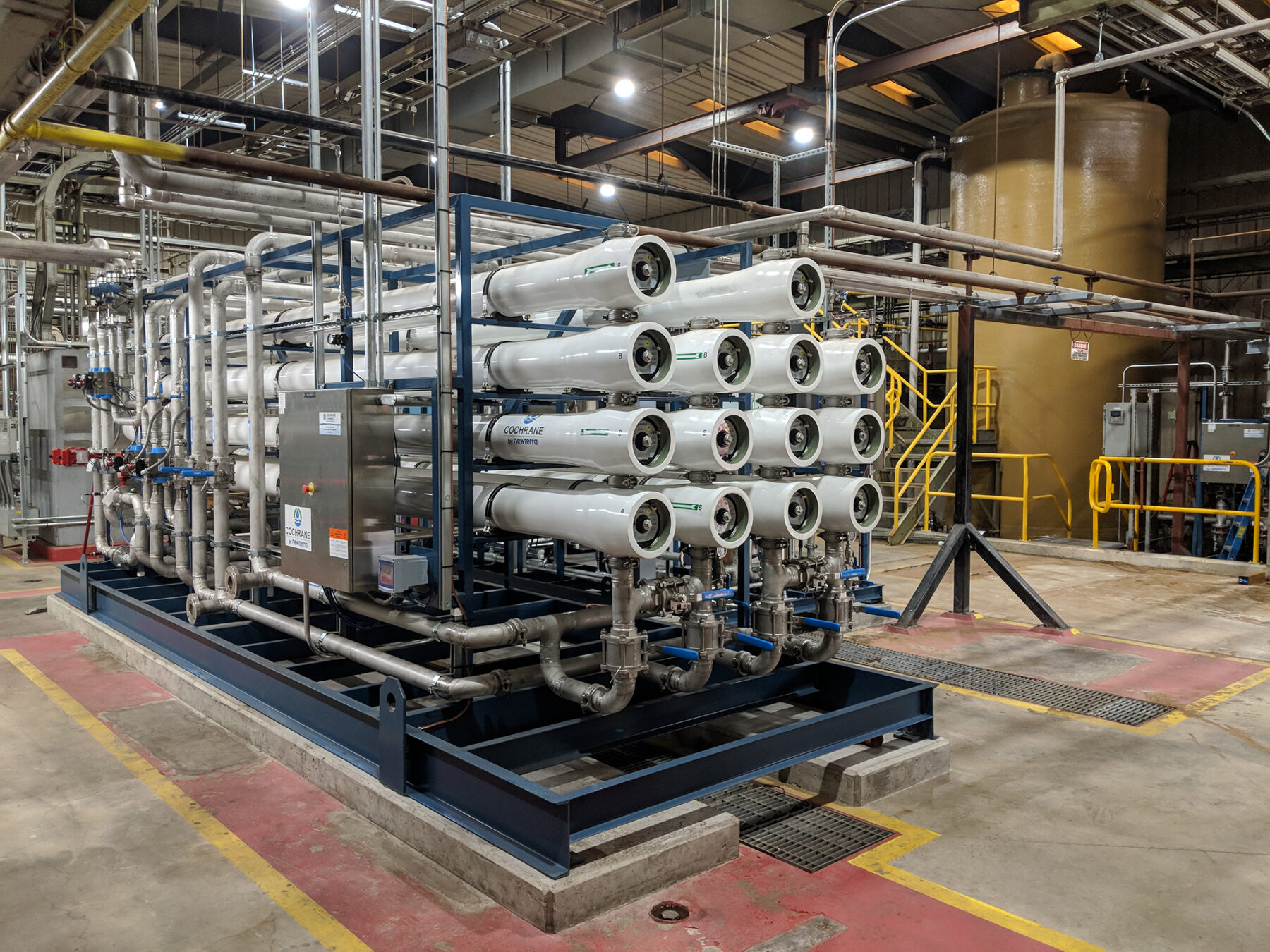 Newterra custom reverse osmosis system consists of a fourteen white pipes stacked in four columns consisting of four pipes, three pipes, four pipes, and three pipes when read left to right with a blue enameled steel frame with steel pipes leading to a control box within a water treatment warehouse.