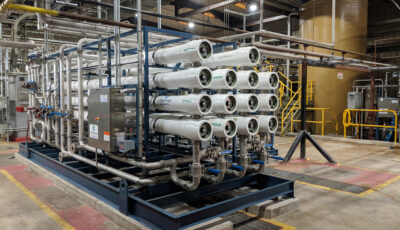 Newterra custom reverse osmosis system consists of a fourteen white pipes stacked in four columns consisting of four pipes, three pipes, four pipes, and three pipes when read left to right with a blue enameled steel frame with steel pipes leading to a control box within a water treatment warehouse.