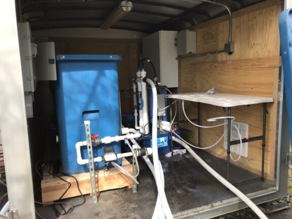 Newterra Stormwaterx technology in use in a modular, portable unit within a shipping container lined with plywood containing a water filtration unit made of blue rigid plastic with white pvc pipes containing valves and connecting to four vertical filters, enclosed electrical panels, and accompanied by a plastic folding table. Four hoses extend outside of the container to the natural environment.