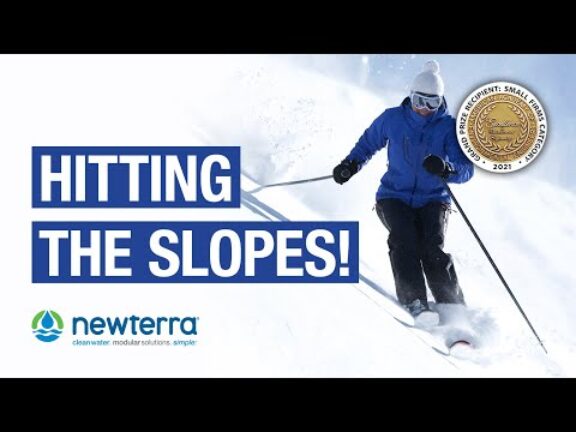 Person in blue jacket and black ski pants and white boots skiing down a snow covered hill with the words 