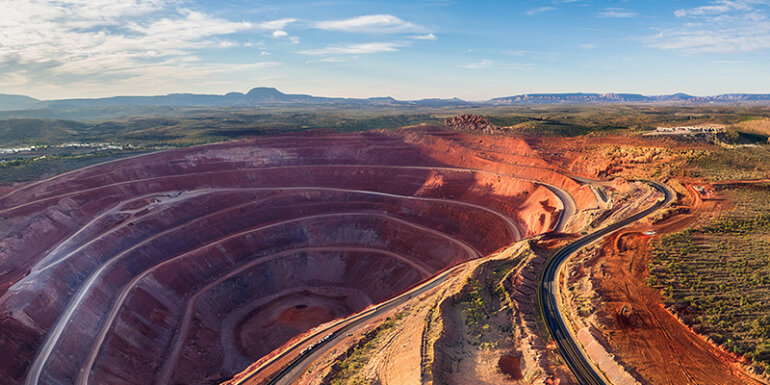 Aerial view of an open-pit rare earth mine in the Southwest, showing terraced red rock excavation levels, winding access roads, and a dry desert landscape stretching to distant mountain ranges under a partly cloudy sky.