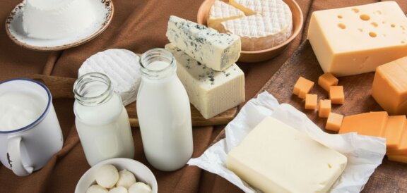 Assorted dairy spread on a wooden board: milk bottles, a mug of milk, yogurt cup, various cheeses and butter blocks/crumbs.