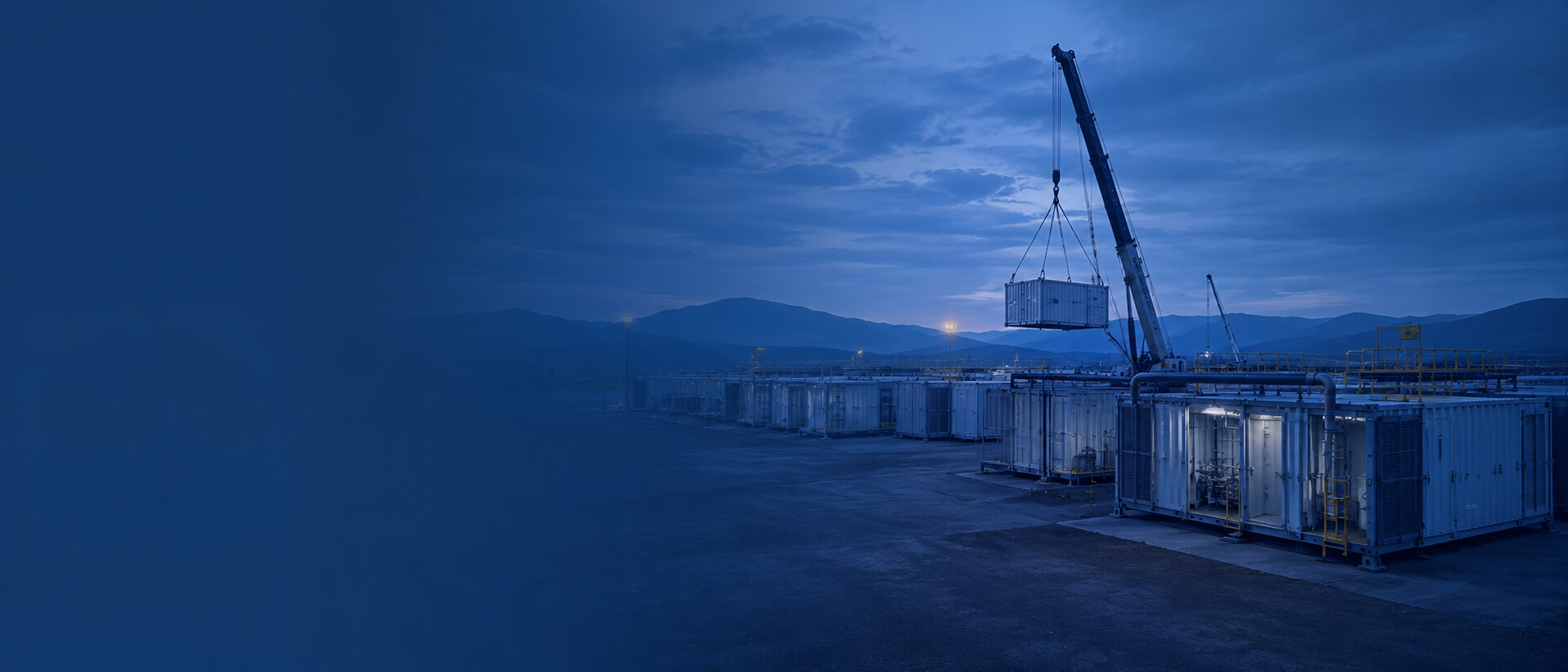 Crane lifting a shipping container over an industrial yard at dusk with blue-tinted lighting