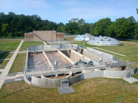 Overhead view of a wastewater treatment plant with circular concrete settling basins, metal railings, and a rectangular building, with green grass and trees in the background and clear blue sky.