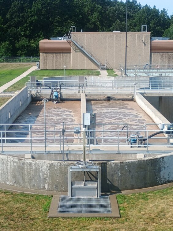 Water treatment plant with circular concrete basins, metal railings, and walkways among grassy areas and trees in the background.